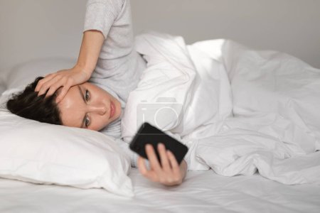 Photo for Sad tired caucasian millennial lady lies on bed, suffers from headache and migraine, reads message on smartphone in bedroom interior. Bad news, health problems, insomnia, stress and negative at home - Royalty Free Image