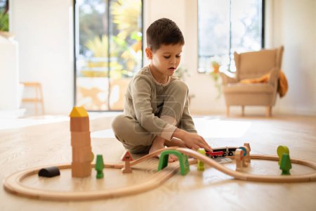Foto de Busy serious caucasian little kid builds city, plays wooden road, train and cars in nursery room interior. Fun alone in kindergarten, entertainment at home, fantasy and childhood, education with game - Imagen libre de derechos
