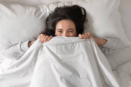 Téléchargez les photos : Happy pretty young european female sleeps on white bed, covers face with blanket, wakes up, enjoys good morning, weekend or free time at vacation in bedroom. Health care, rest and relaxation at home - en image libre de droit