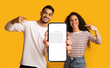 Téléchargez les photos : Place For Ad. Cheerful Middle Eastern Couple Demonstrating Big Blank Smartphone With White Screen For Mockup And Pointing At It, Happy Arab Man And Woman Advertising New Mobile App Or Website - en image libre de droit