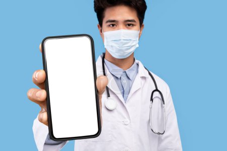 Foto de Asian Doctor Wearing Protective Mask Showing Big Smartphone With Blank White Screen, Young Chinese Male Physician Holding Cellphone With Copy Space For Mobile Advertisement, Collage, Mockup - Imagen libre de derechos