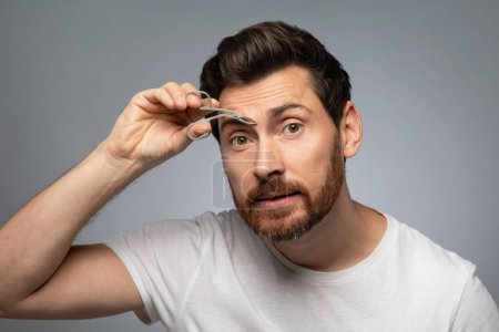 Photo for Grooming wellness and facial care. Handsome middle aged bearded man tweezing eyebrows, standing over grey background, studio shot - Royalty Free Image