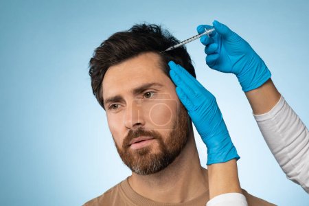 Mesotherapie für Haare. Mann mittleren Alters bekommt Injektionen in Kopf, Mesotherapie, Therapeut in Schutzhandschuhen mit Spritze, blauer Studiohintergrund