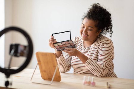 Téléchargez les photos : Cheerful pretty curly young chubby hispanic lady beauty blogger sitting at desk full of various makeup products, recording video on smartphone, showing eyeshadow palette, recommending cosmetics - en image libre de droit
