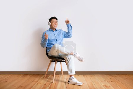 Téléchargez les photos : Excited middle aged asian man listening music in wireless stereo headphones and dancing, sitting on chair over white background wall, copy space, full length - en image libre de droit