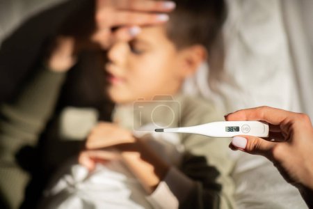 Foto de Unhappy caucasian little child suffers from fever, mom measures temperature, show thermometer, touch forehead in bedroom, cropped. Flu and cold, health care, childhood, illness, treatment at home - Imagen libre de derechos