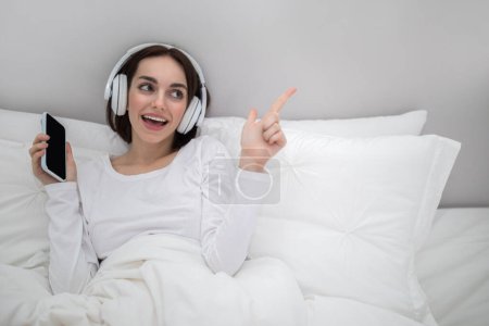 Téléchargez les photos : Cute happy young brunette lady wearing white pajamas listening to music, singing karaoke and dancing in bed at home, using smartphone with mockup and newest wireless headphones, pointing at copy space - en image libre de droit