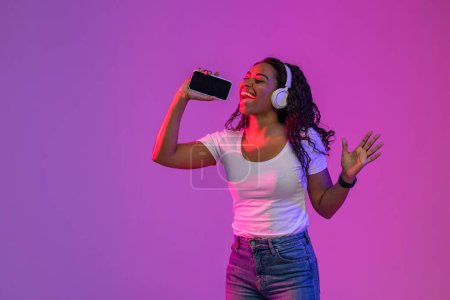Téléchargez les photos : Enthousiaste afro-américaine femme écoutant de la musique dans les écouteurs via App sur Smartphone, positive jeune femme noire en utilisant le téléphone mobile comme microphone tout en se tenant debout dans la lumière au néon sur fond violet - en image libre de droit