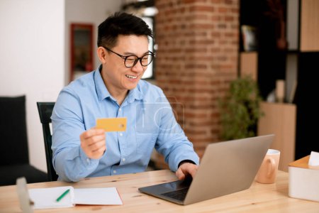 Photo for Fast online shopping. Happy middle aged asian businessman holding debit credit card and using laptop, making financial transaction while sitting at table at home office, copy space - Royalty Free Image