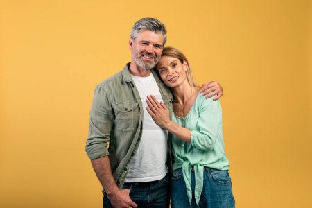Téléchargez les photos : Portrait of happy mature couple hugging and posing at camera, cheerful middle aged spouses standing over yellow studio background, smiling together, copy space - en image libre de droit