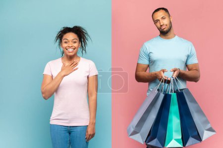 Téléchargez les photos : Homme noir mécontent debout avec des sacs à provisions, faisant du shopping avec sa dame ravie, debout sur fond de studio rose et bleu. Shopaholism, concept de couple incompatible - en image libre de droit