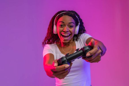 Téléchargez les photos : Jeune femme noire excitée avec joystick jouant à des jeux vidéo dans l'éclairage au néon, femme africaine joyeuse portant des écouteurs s'amusant, jouissant de jeux virtuels tout en se tenant debout sur fond violet - en image libre de droit
