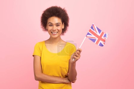 Téléchargez les photos : Cheerful pretty millennial multiethnic woman in casual hold England flag, enjoy tutoring, isolated on pink background, studio. Exchange study, recommendation for education, learn English, ad and offer - en image libre de droit