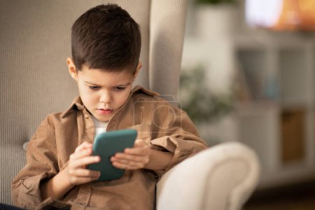 Photo for Busy serious pretty caucasian little child with smartphone sits in armchair, watches videos in living room interior, close up. Game and learning app, study and gadget addiction at spare time at home - Royalty Free Image