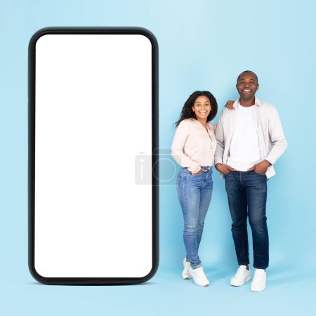 Foto de Happy african american spouses standing near large cellphone with blank screen, advertising application or website, posing over blue studio background. Full length, mockup - Imagen libre de derechos