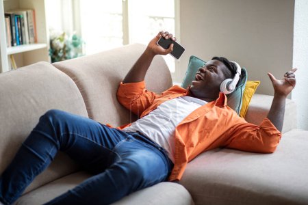 Téléchargez les photos : Emotional happy stylish chubby millennial black guy in casual lying on couch in living room, using wireless headphones and cell phone, singing songs at home, karaoke mobile app, copy space - en image libre de droit