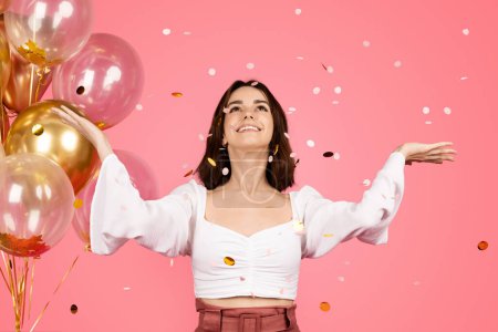 Téléchargez les photos : Satisfied inspired pretty young european lady with many balloons enjoying confetti, has fun isolated on pink background, studio. Great mood, win, celebration holiday, birthday, party ad and offer - en image libre de droit