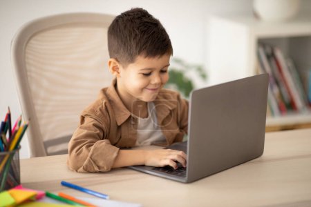 Photo for Video call remotely, education game. Glad caucasian little child pupil sits at table, typing on computer, play, watch video in room interior. Study at school, kindergarten and home, childhood and app - Royalty Free Image