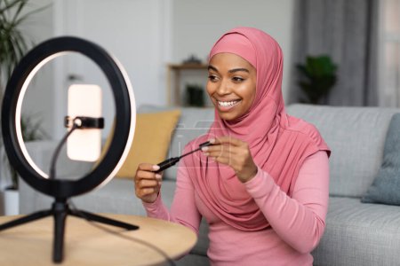 Foto de Beauty blogger. Happy african american lady in hijab reviewing new mascara, looking at smartphone set on ring lamp, recording video for her blog - Imagen libre de derechos