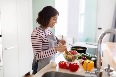 Téléchargez les photos : Cooking, culinary, domestic lifestyle, chores concept. Smiling cheerful happy pretty young arab woman housewife preparing dinner at home, mixing fresh organic vegetables, side view, copy space - en image libre de droit