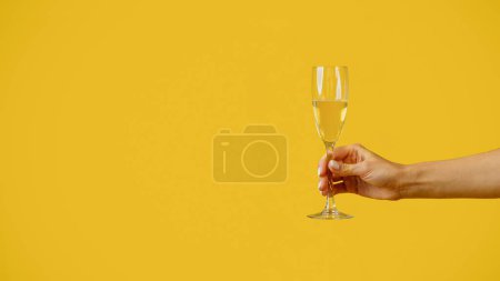 Foto de Female hand holding glass of champagne on yellow studio background, panorama with free copy space. Unrecognizable lady having fun party, celebrating holiday with alcoholic drink - Imagen libre de derechos