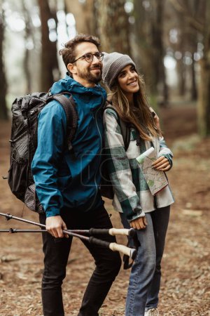 Téléchargez les photos : Souriant jeune couple européen touristes en vestes avec sac à dos, bâtons de trekking et carte marcher dans la forêt, profiter de la saison froide en plein air, regarder l'espace libre. Aventure et relations, mode de vie, voyage - en image libre de droit