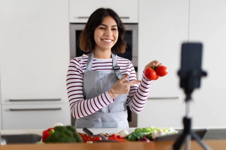 Photo for Positive pretty young middle eastern woman wearing apron influencer or food blogger streaming while cooking at home, showing bunch of fresh juicy tomatoes at smartphone camera, kitchen interior - Royalty Free Image