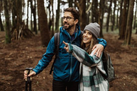 Glad young european couple tourists in jackets with backpack and trekking sticks walk in forest, point finger at free space, enjoy cold season outdoor. Active lifestyle travel, adventure and weekend