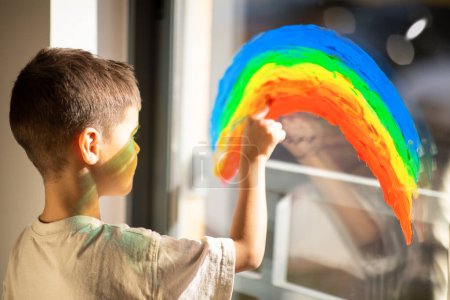 Foto de Caucasian little child draws rainbow with paint on window in elementary school, kindergarten room interior, close up. Fun alone, entertainment and play at home, art, education, fantasy and childhood - Imagen libre de derechos