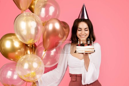 Photo for Happy pretty young european woman in hat holding many balloons and cake with candles, makes a wish, has fun, isolated on pink background, studio. Celebration holiday, birthday, dream and anniversary - Royalty Free Image