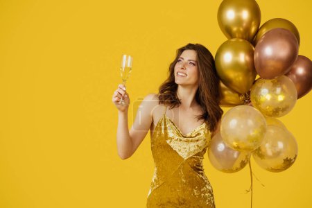 Photo for Beauty lady in dress holding air balloons and glass of champagne, looking at free space on yellow background, studio shot. Happy woman on birthday holiday party having fun - Royalty Free Image