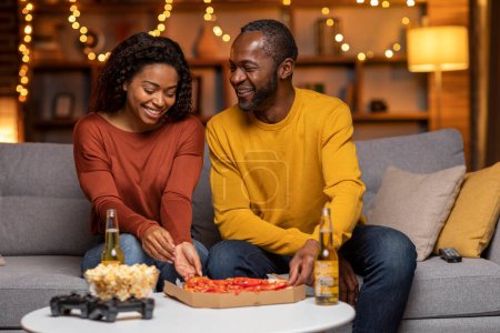 Beautiful happy loving african american middle aged man and millennial woman sitting on couch, having conversation, drinking beer, eating pizza, black couple enjoying nice evening at home