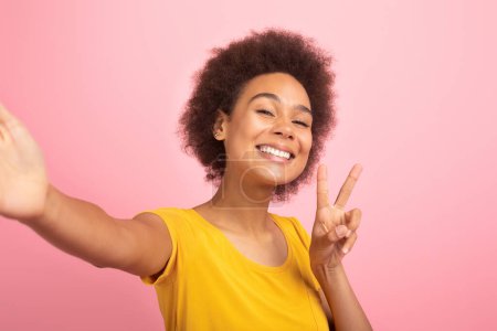 Photo for Glad excited millennial multiethnic woman in casual taking selfie and show peace sign with hand isolated on pink background, studio, close up. Photo for blog and social networks, good ad and offer - Royalty Free Image