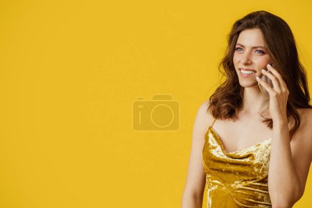 Photo for Portrait of pretty lady talking on cellphone and smiling, standing isolated on yellow studio background, talking and looking away at free space. Remote communication - Royalty Free Image