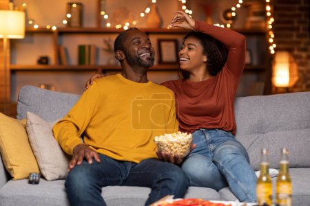 Téléchargez les photos : Playful happy beautiful adult african american lovers man and woman in casual having fun at cozy house in the evening, cheerful wife feeding her laughing husband with popcorn, copy space - en image libre de droit