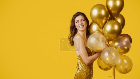 Foto de Hora de la fiesta. Encantadora mujer feliz en vestido sosteniendo racimo de globos sobre fondo de estudio amarillo, panorama con espacio de copia. Jovencita divirtiéndose celebración, disfrutando vacaciones - Imagen libre de derechos
