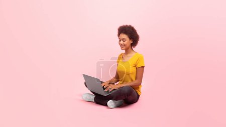Foto de Cheerful millennial multiethnic lady typing on computer sits on floor and chatting, isolated on pink background, studio, panorama, free space. Gadget for study, work, social networks, ad and offer - Imagen libre de derechos