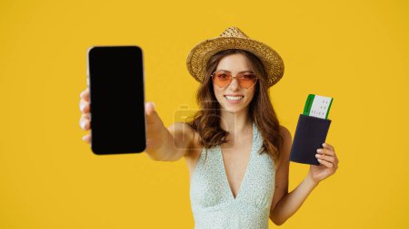 Photo for Booking app. Happy lady showing blank smartphone and holding passport with tickets, standing over yellow background, ready for vacation trip, mockup - Royalty Free Image