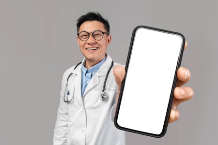 Photo for Asian male doctor showing smartphone with big blank screen at camera, smiling chinese medical worker in uniform demonstrating mobile phone with copy space for advertisement, collage, mockup - Royalty Free Image