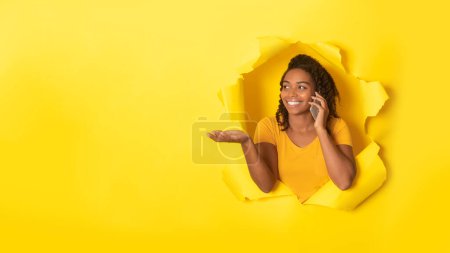 Téléchargez les photos : Excited black lady talking on cellphone and looking aside at free space, posing in torn yellow paper background, panorama. Mobile offer, communication concept - en image libre de droit
