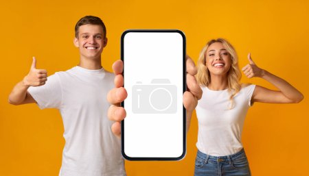 Photo for Smiling Young Couple Holding Big Blank Smartphone In Hand And Showing Thumb Up Gesture, Happy Man And Woman Recommending New Mobile Application Or Website, Standing On Yellow Background, Mockup - Royalty Free Image