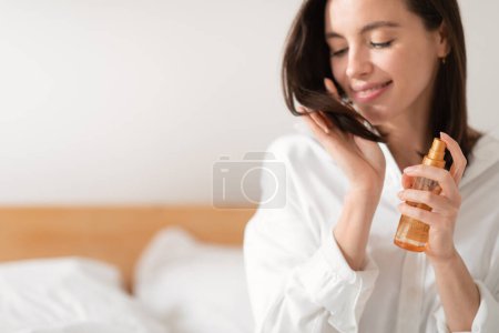 Photo for Glad caucasian millennial female sits on bed puts spray on hair, looks at ends, enjoy good morning, treatment in bedroom interior. Ad and offer, beauty care, routine procedures in spare time at home - Royalty Free Image