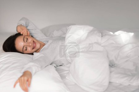 Photo for Glad young european lady sleeping on white bed with closed eyes, waking up, stretching body, enjoying good morning, weekend or vacation in bedroom interior. Health care, rest and relaxation at home - Royalty Free Image