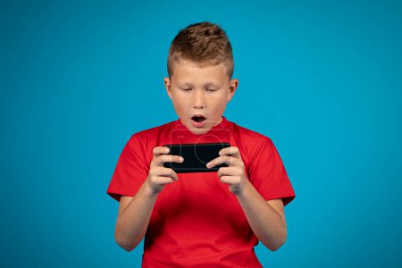 Photo for Shocked preteen boy playing on mobile phone on blue studio background, cute male kid looking at smartphone screen and opening mouth in amazement, enjoying online game, having gadget addiction - Royalty Free Image