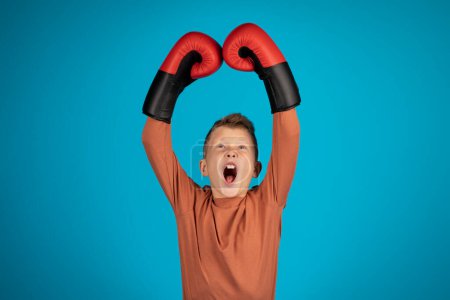 Téléchargez les photos : Portrait Of Agressive Preteen Boy Wearing Boxing Gloves Raising Arms And Shouting, Sporty Male Child Emotionally Celebrating Win While Standing Isolated Over Blue Background, Copy Space - en image libre de droit