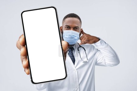 Téléchargez les photos : Online Consultation. Black Male Doctor Wearing Mask Holding Big Blank Smartphone And Gesturing Call Me, Male Physician Showing Cellphone With Copy Space For Mobile Advertisement, Collage, Mockup - en image libre de droit