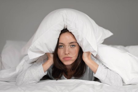 Photo for Unhappy tired young european woman lies on white comfortable bed under blanket, covers her head with pillow, wakes up in bedroom. Problems with rest and relax at home, insomnia and noisy neighbors - Royalty Free Image