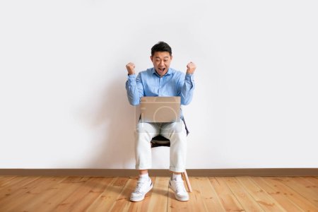 Photo for Overjoyed middle aged asian man with laptop computer sitting on chair, celebrating great deal or business success, making YES gesture against white studio wall, copy space, full length - Royalty Free Image