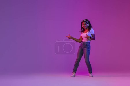 Téléchargez les photos : Joyful African American Female In Wireless Headphones Pointing Aside At Copy Space, Positive Young Black Woman Standing In Neon Light, Listening Music And Showing Free Place For Advertisement - en image libre de droit