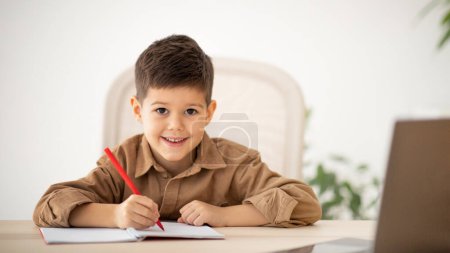 Photo for Video call, study remotely. Smiling caucasian little child sit at table, making notes, watching lesson on computer in room interior. Education at school, kindergarten and home, childhood and lifestyle - Royalty Free Image